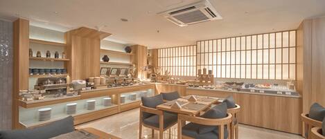 Una cafetería moderna y bien iluminada, con mobiliario de madera, una barra central y un gran ventanal con persianas verticales.