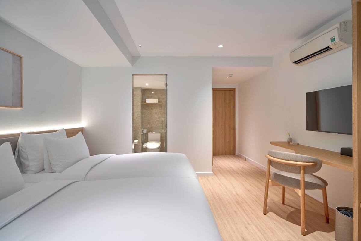 A modern hotel room with a large bed, a desk with a chair, a TV, and an air conditioning unit.