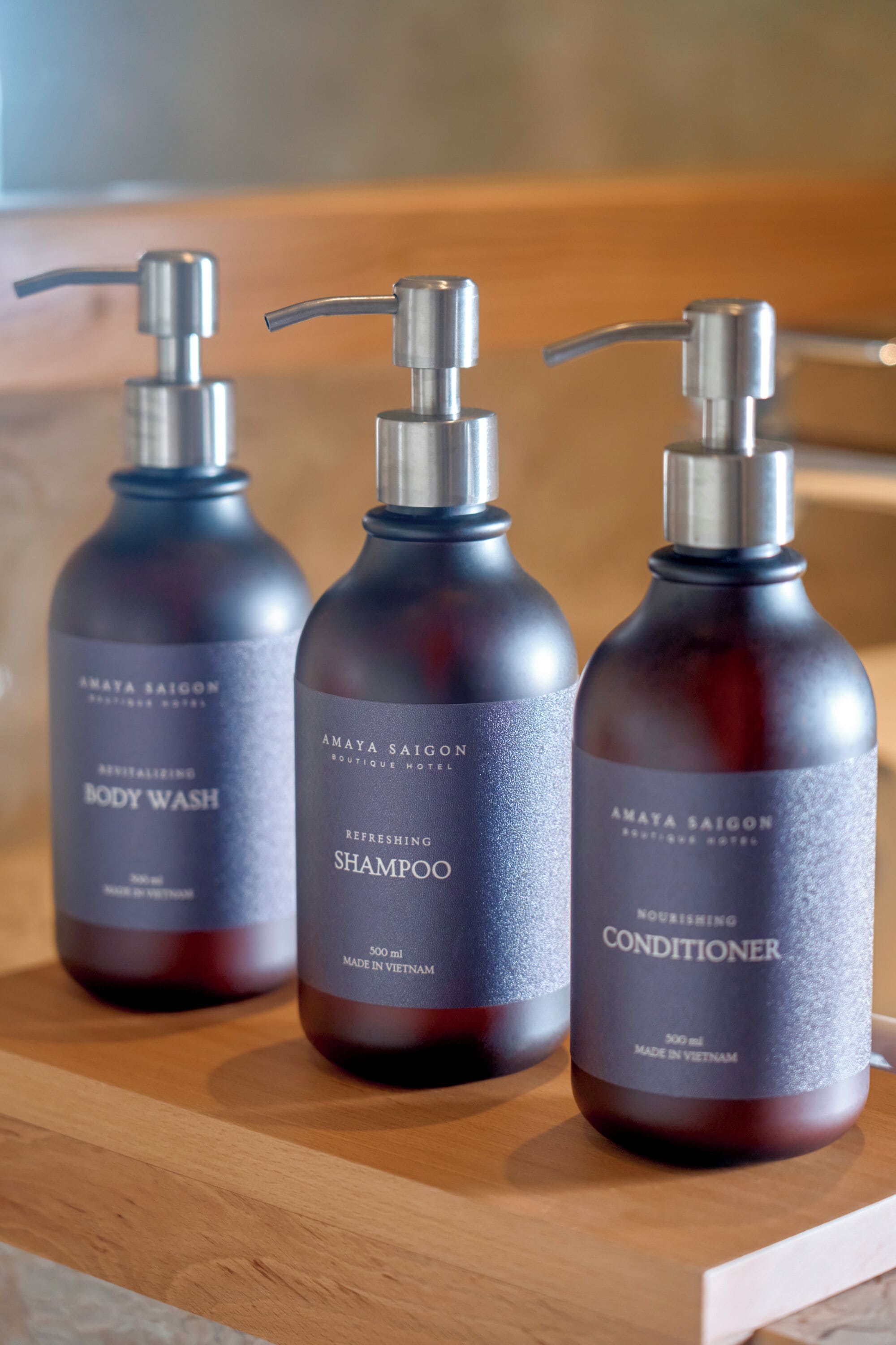 Tres botellas de gel de baño, champú y acondicionador Amaya Saigon sobre una superficie de madera.