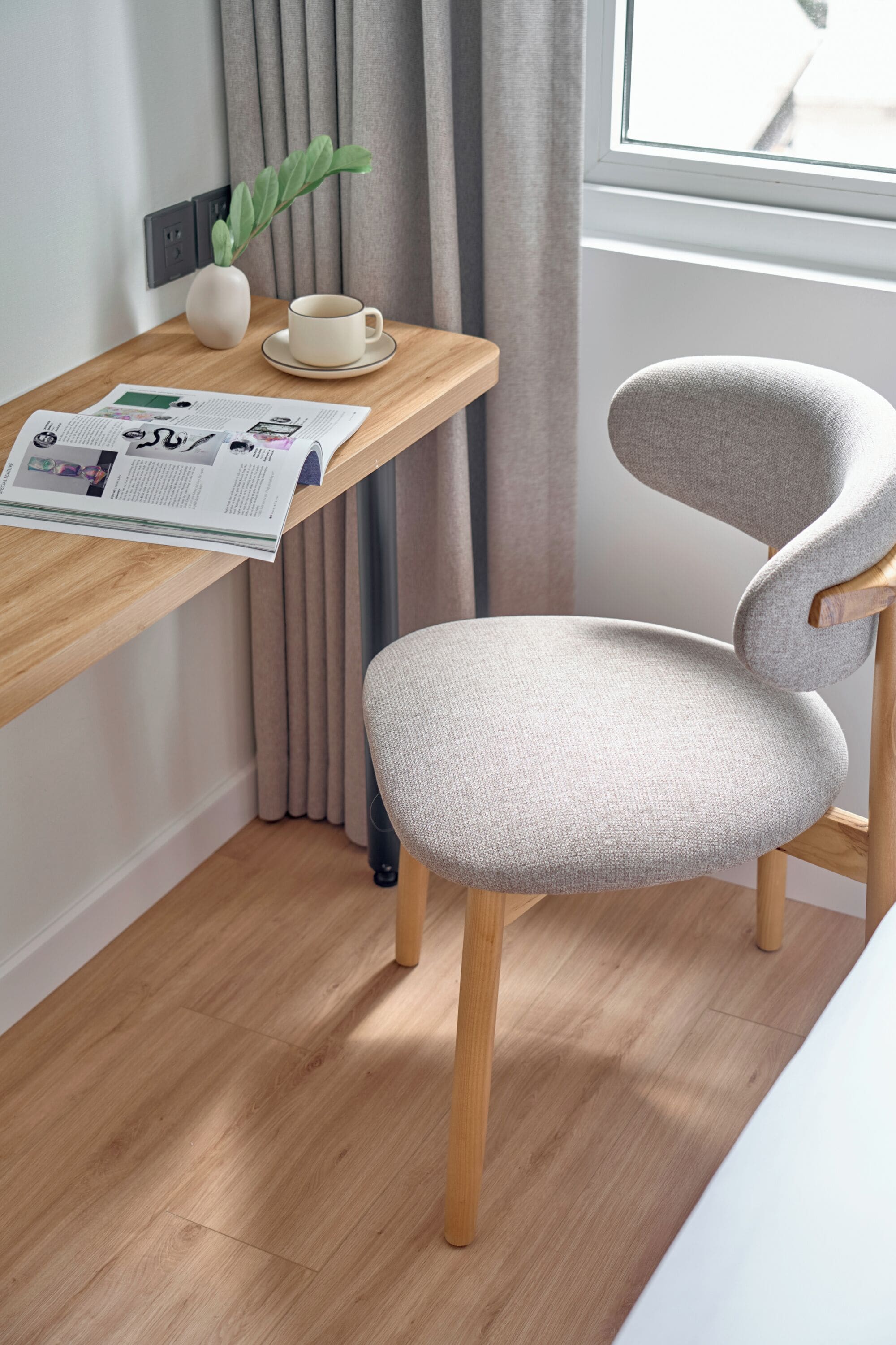 Una habitación moderna con un escritorio de madera, una silla tapizada de gris, una taza blanca, un florero con una planta y un periódico.