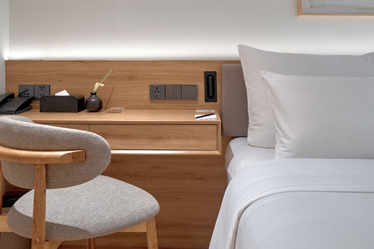 A hotel room with a wooden desk, a chair, a bed, and a bedside table with a lamp.