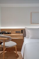 A hotel room with a wooden desk, a chair, a bed, and a bedside table with a lamp.