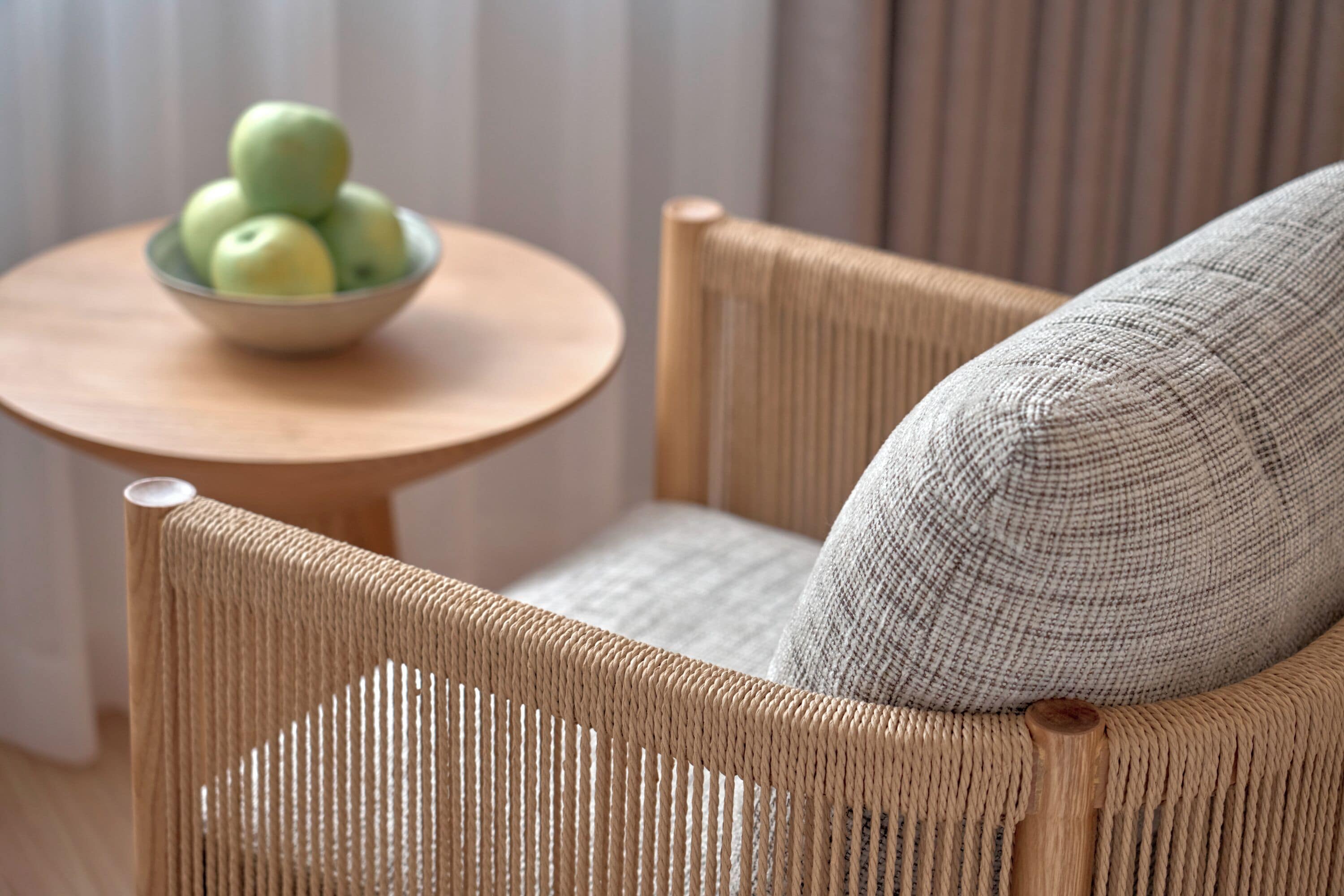 A wicker chair with a cushion, a round wooden table with a bowl of green apples, and a striped curtain in the background.