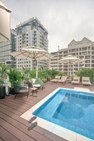 Un área de piscina en la azotea con sillones de descanso y sombrillas, rodeada de plantas en macetas y vistas del edificio del hotel The Lancaster.