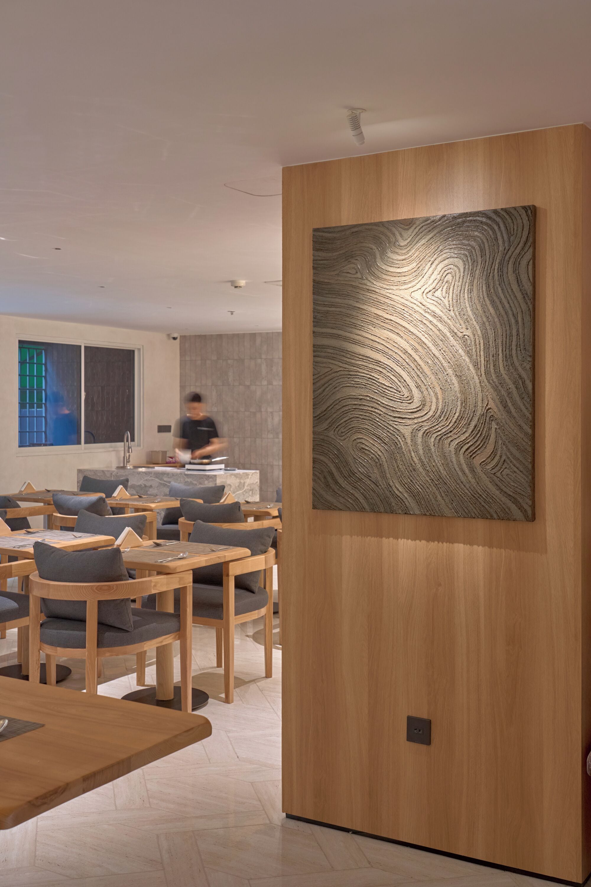 A modern dining area with wooden tables and chairs, a large abstract painting on the wall, and a kitchen in the background.
