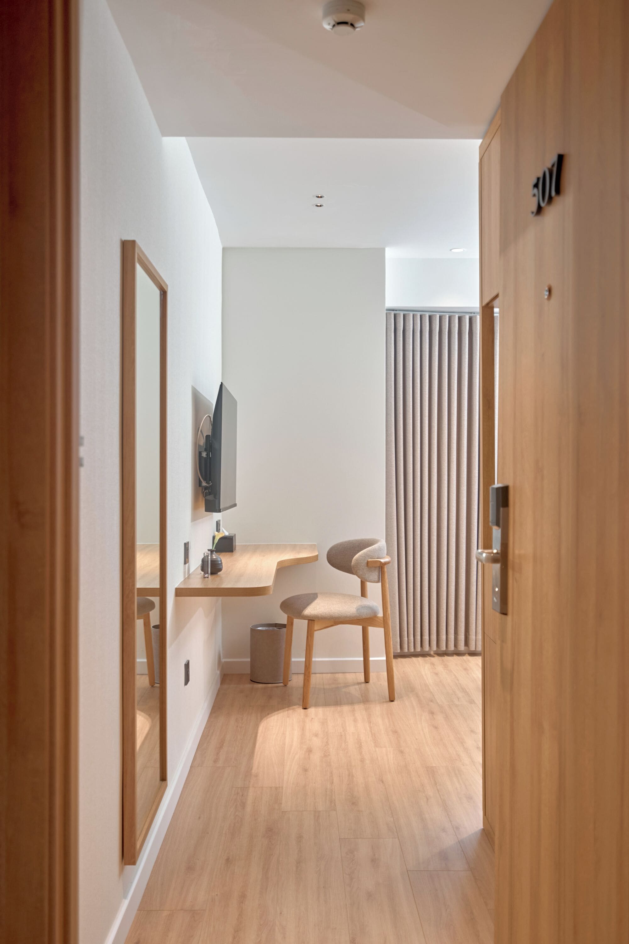 A modern hotel room with a wooden floor, a desk with a chair, a television, and a door marked with the number 501.