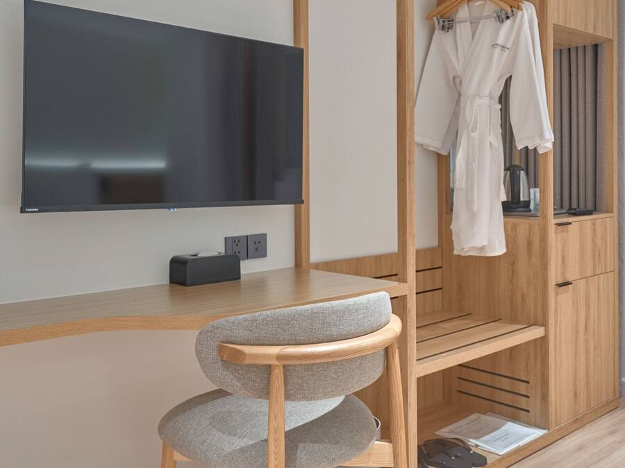Una habitación moderna con un armario de madera, una butaca tapizada de gris, un televisor de pantalla plana y una bata colgada.