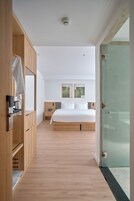 Quarto de hotel moderno com guarda-roupa de madeira, cama com lençóis brancos, mesa de cabeceira com luminária e porta de vidro que dá para uma varanda.