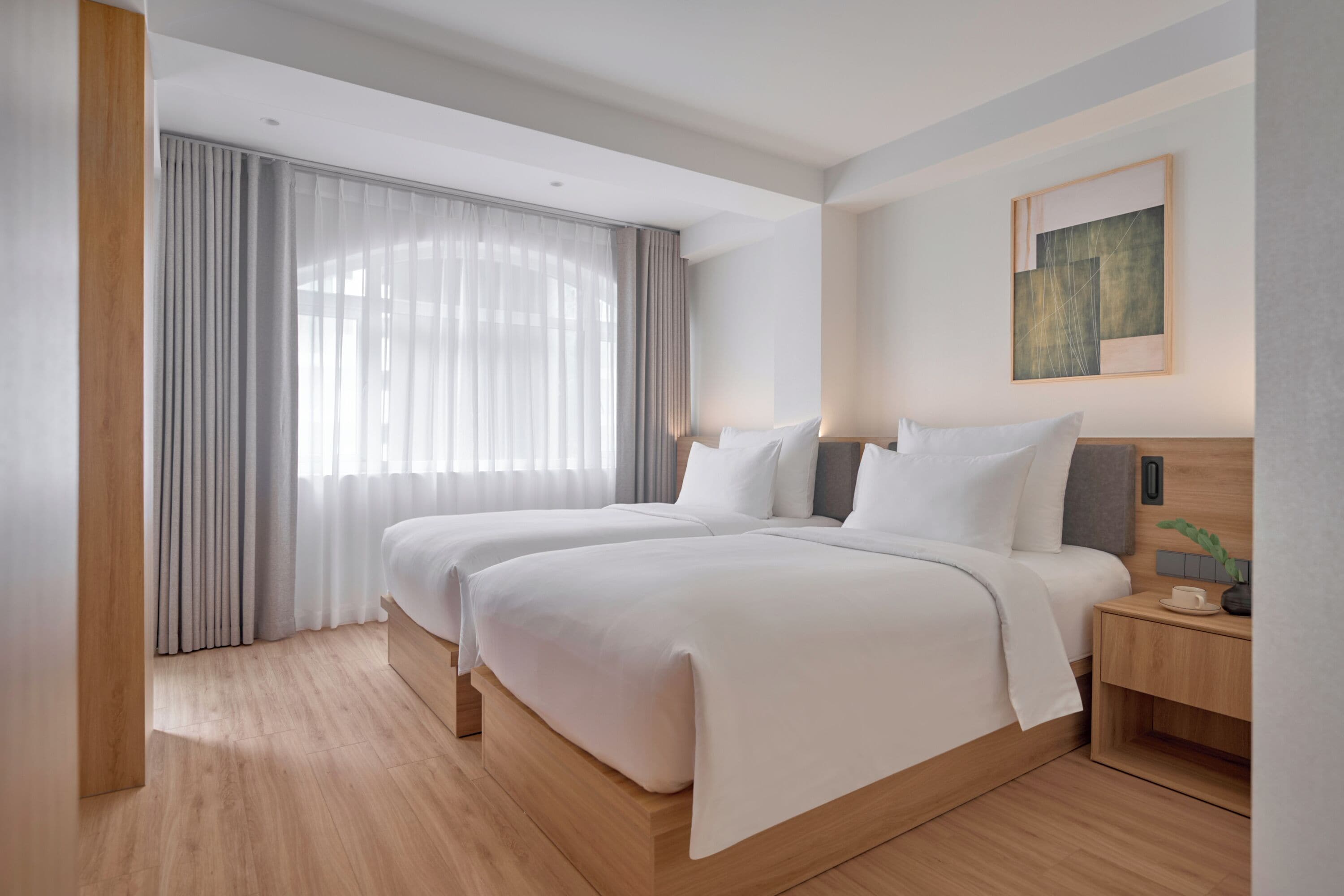 A modern hotel room with two beds, a large window with sheer curtains, a wooden floor, and a minimalist decor.