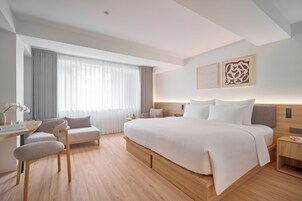 A modern hotel room with a large bed, a desk, a chair, and a window with curtains.