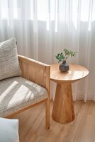 A room with a woven chair, a round wooden table with a vase, and sheer curtains.
