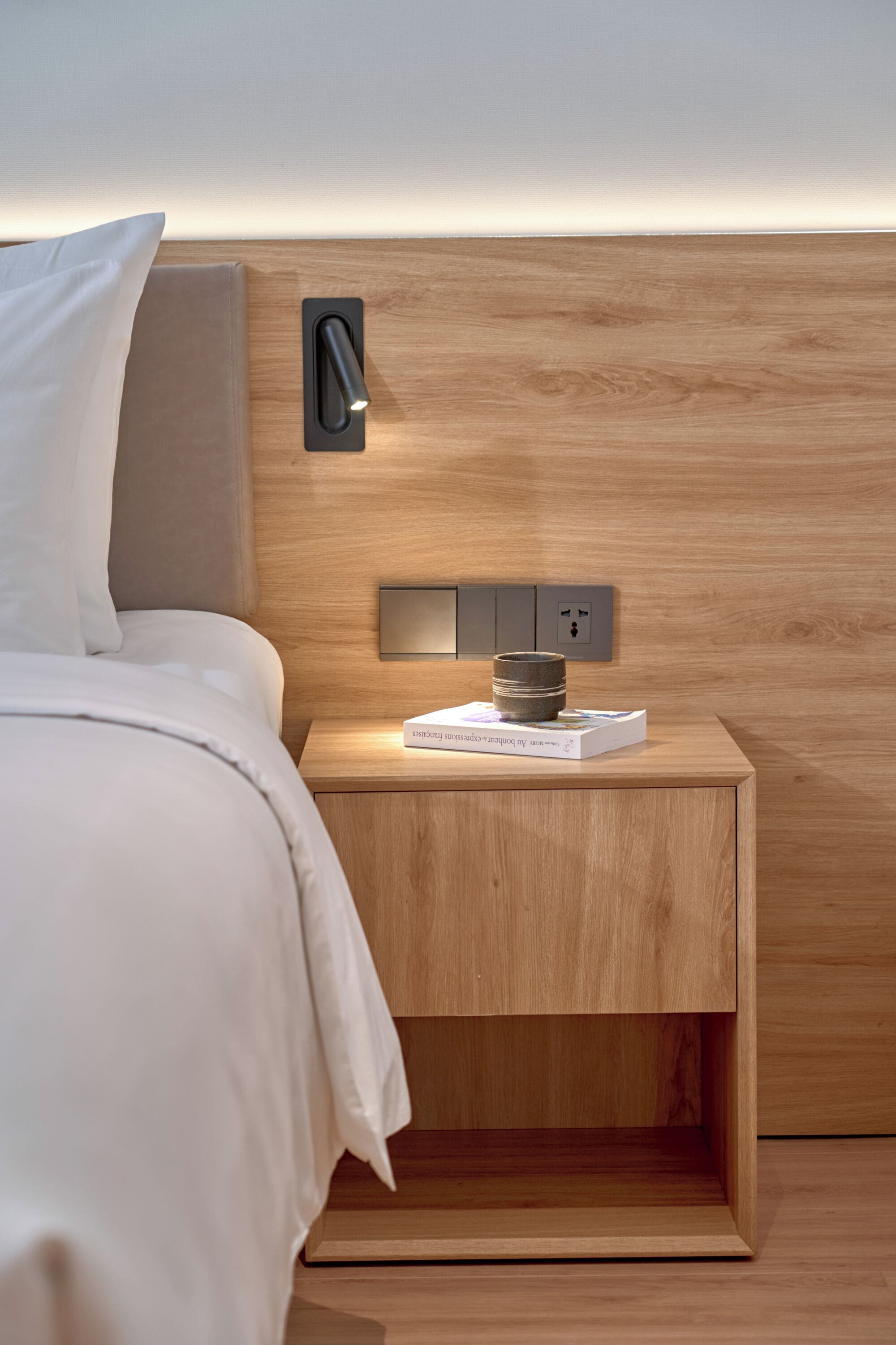 A neatly made bed with a white comforter and pillows, a wooden nightstand with a book and a small container, and a wall-mounted light fixture.
