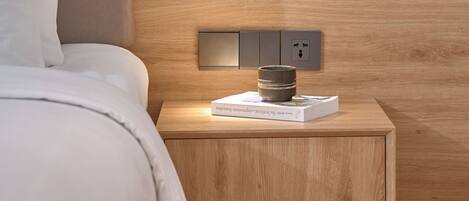 A neatly made bed with a white comforter and pillows, a wooden nightstand with a book and a small container, and a wall-mounted light fixture.