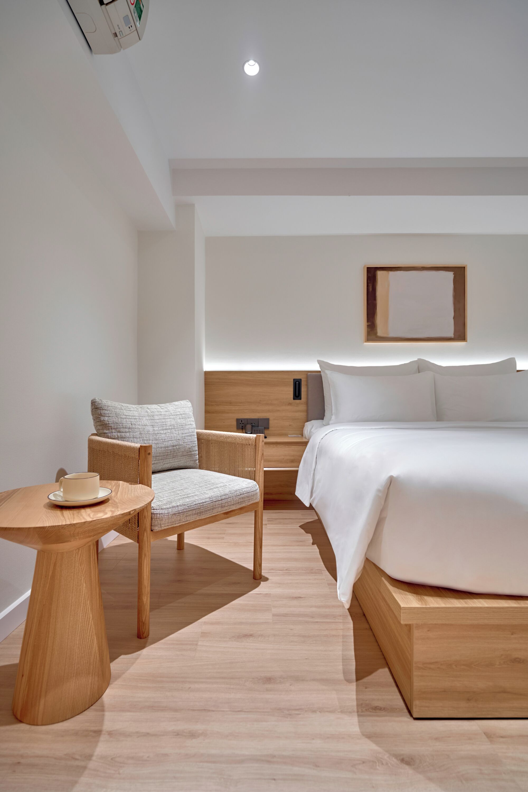 Una habitación de hotel moderna con una cama de madera, una mesa redonda de madera, una silla de mimbre y una lámpara fijada a la pared.