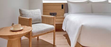 A modern hotel room with a wooden bed, a round wooden table, a wicker chair, and a wall-mounted light fixture.