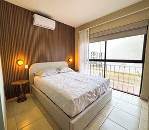 Une chambre à coucher avec un lit, deux lampes de chevet, une petite table et une fenêtre donnant sur des immeubles.