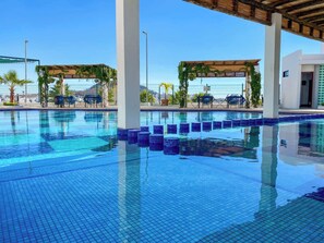 Une piscine aux eaux cristallines, bordée de carreaux, entourée de chaises longues et de parasols, avec en arrière-plan la vue d’un bâtiment moderne.