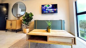 Un salon moderne avec une table basse en bois, un grand miroir, un téléviseur fixé au mur et une plante en pot.