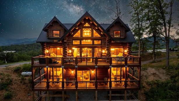 Une cabane en rondins à plusieurs niveaux, dotée de grandes fenêtres et d’un balcon, et entourée d’un paysage boisé.