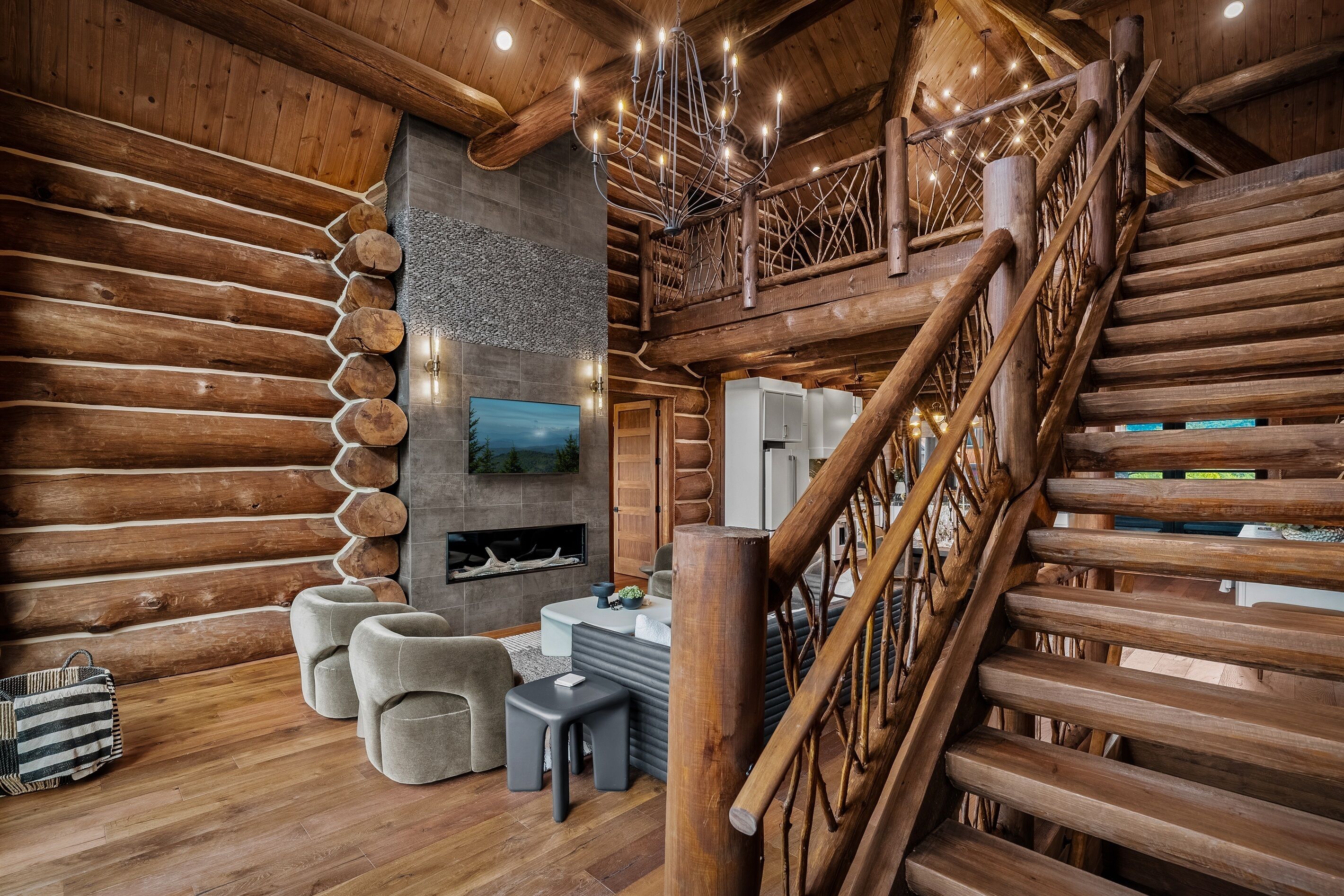 Interior de una cabaña de troncos con escalera de madera, chimenea y televisor colgado en la pared.