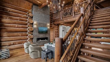 Interior de una cabaña de troncos con escalera de madera, chimenea y televisor colgado en la pared.