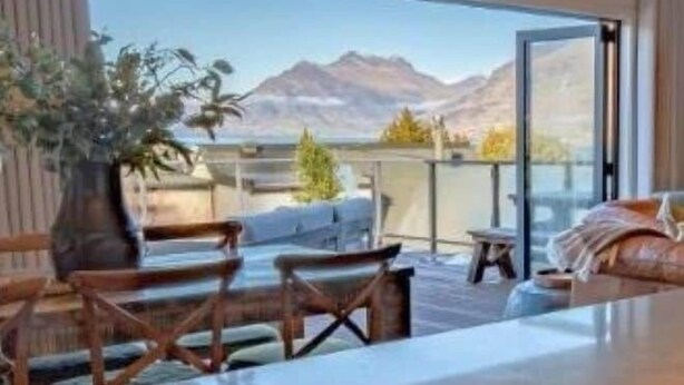 Ett modernt rum med utsikt över bergen, en skylt med texten ”QUEENSTOWN” och en sittgrupp med ett bord och stolar.