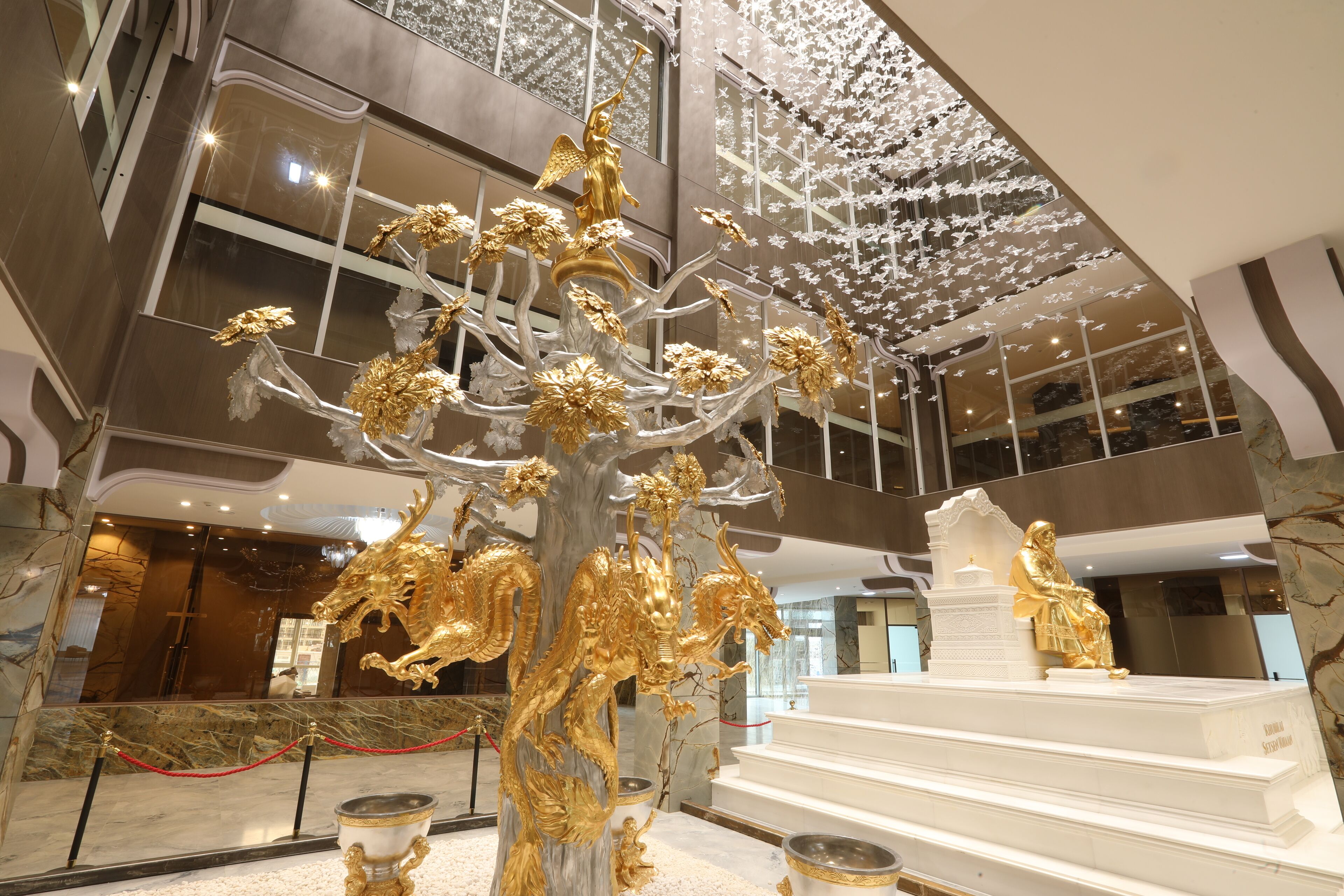 A grand lobby with a large golden tree sculpture, a statue of a seated figure, and a modern chandelier.