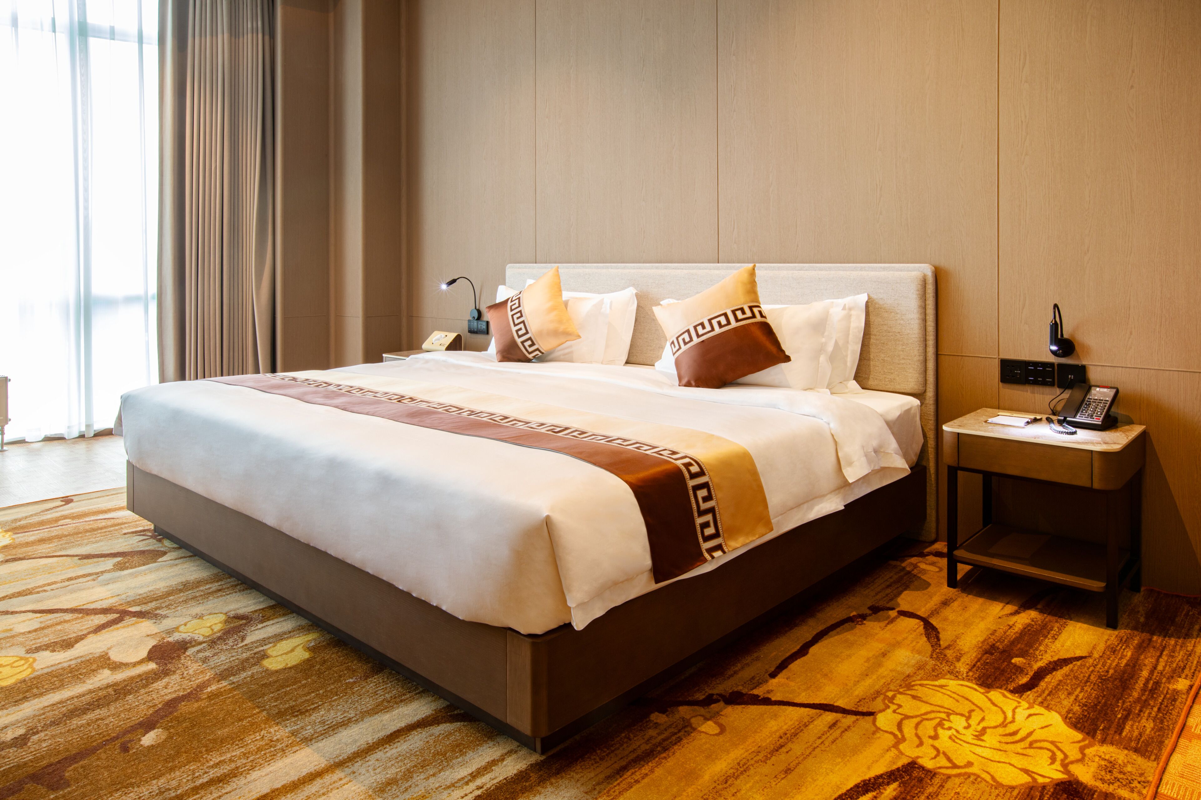 A neatly made bed with a patterned bedspread, a bedside table with a phone and lamp, and a carpeted floor.
