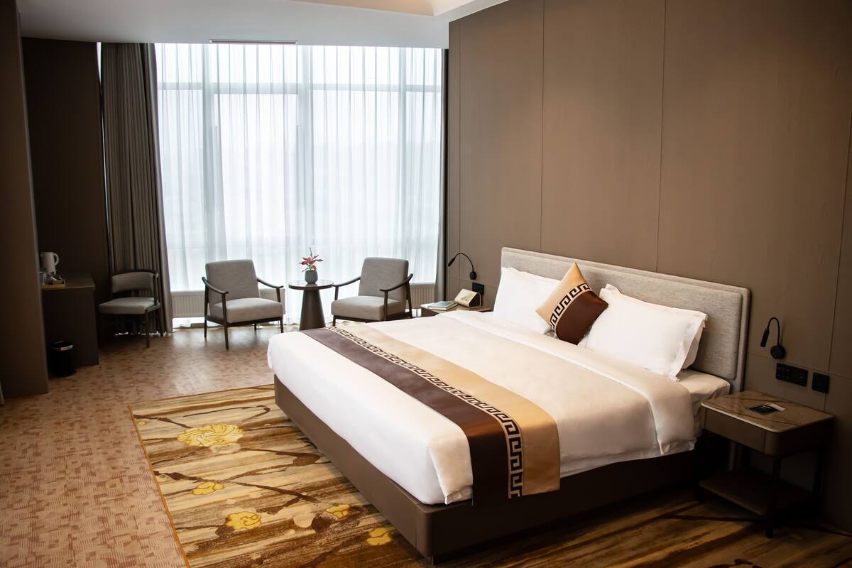 A hotel room with a large bed, two armchairs, a small table, a lamp, and a view of the outdoors through large windows.