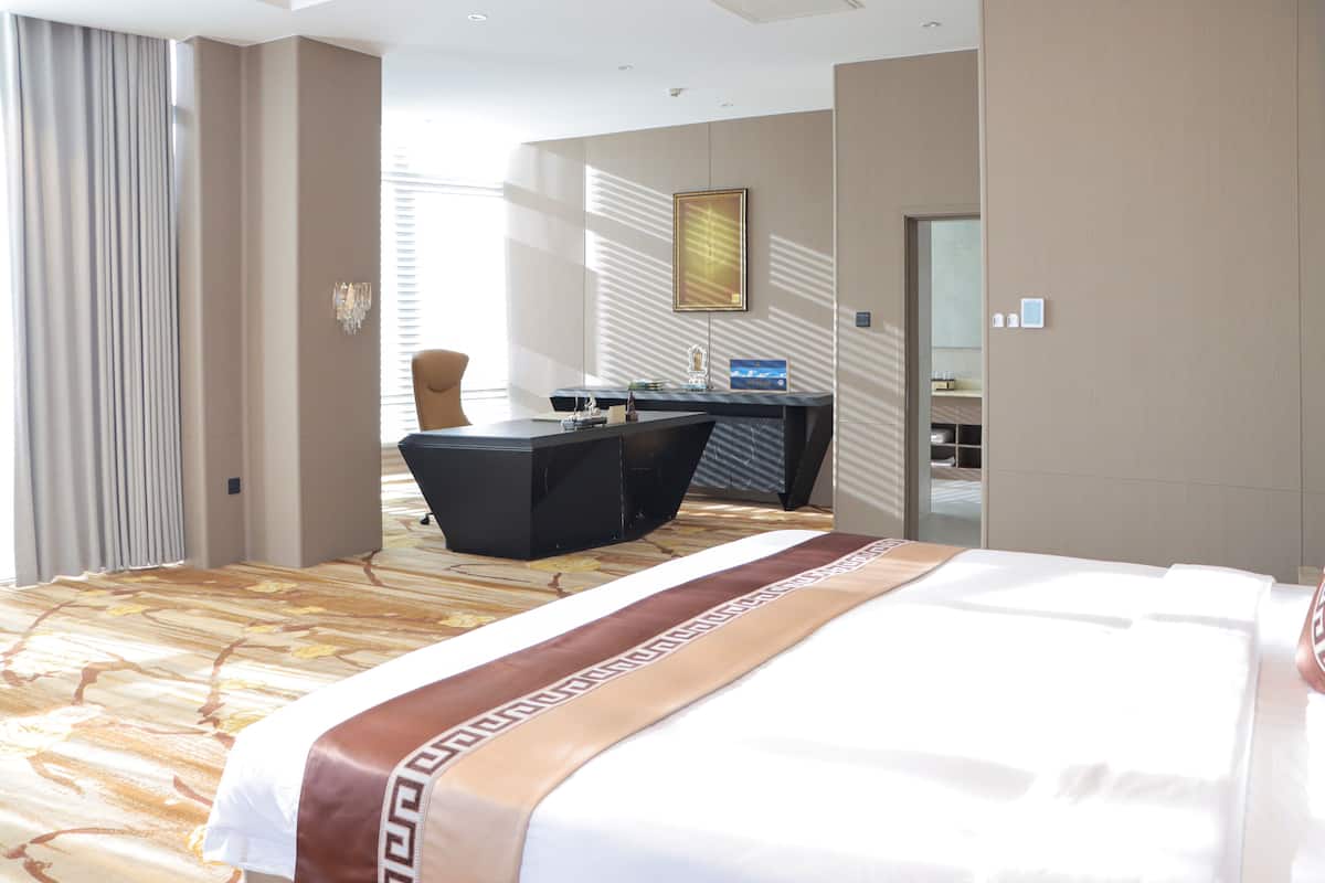 A hotel room with a large bed, a desk, and a chair. There is a window with curtains and a painting on the wall.