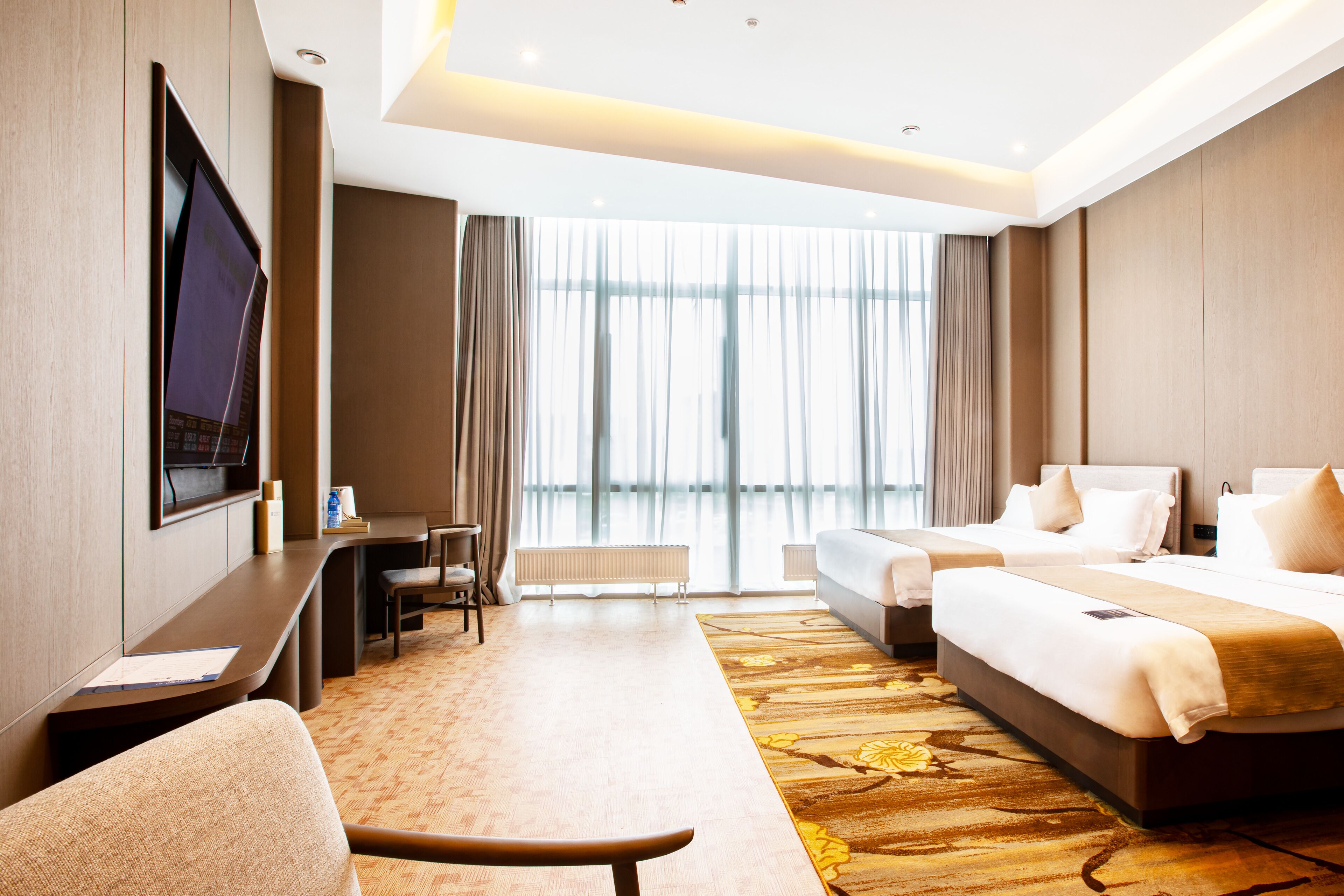 A hotel room with a large bed, a desk, a chair, a TV mounted on the wall, and a large window with curtains.