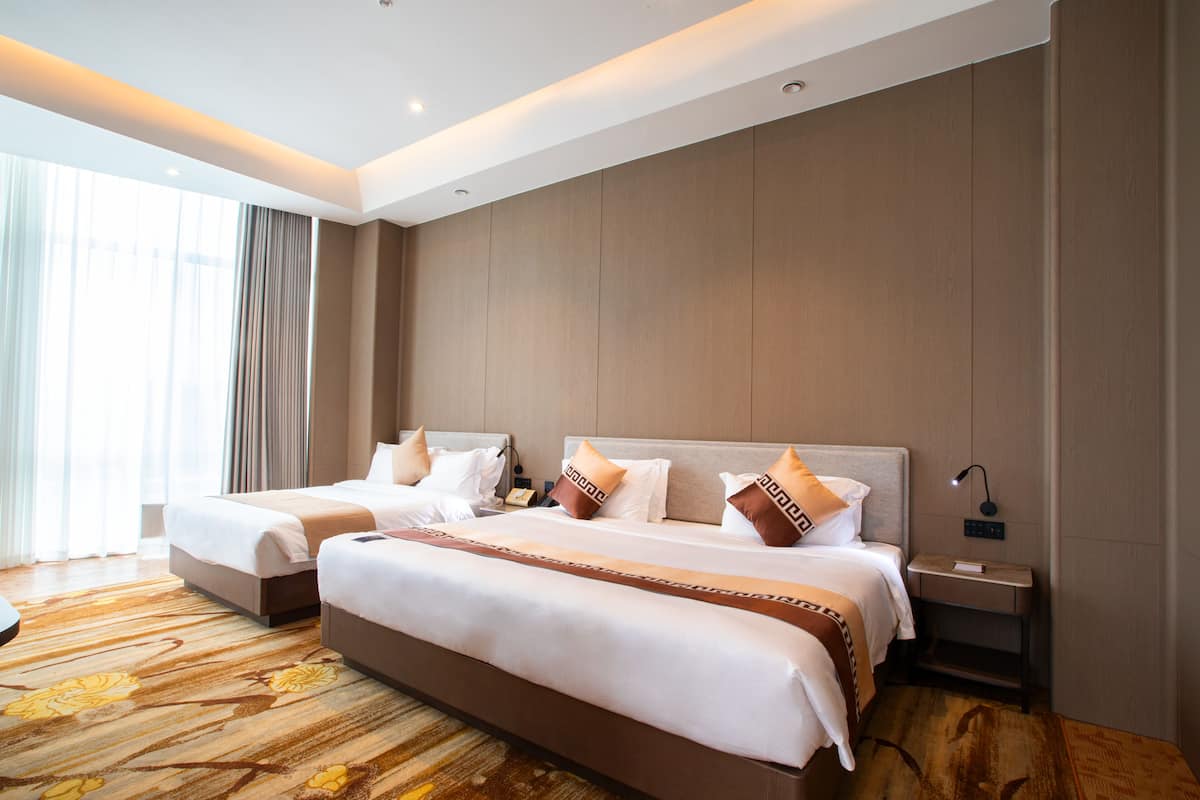 A hotel room with two beds, a large window with curtains, a wooden floor, and a nightstand with a lamp.
