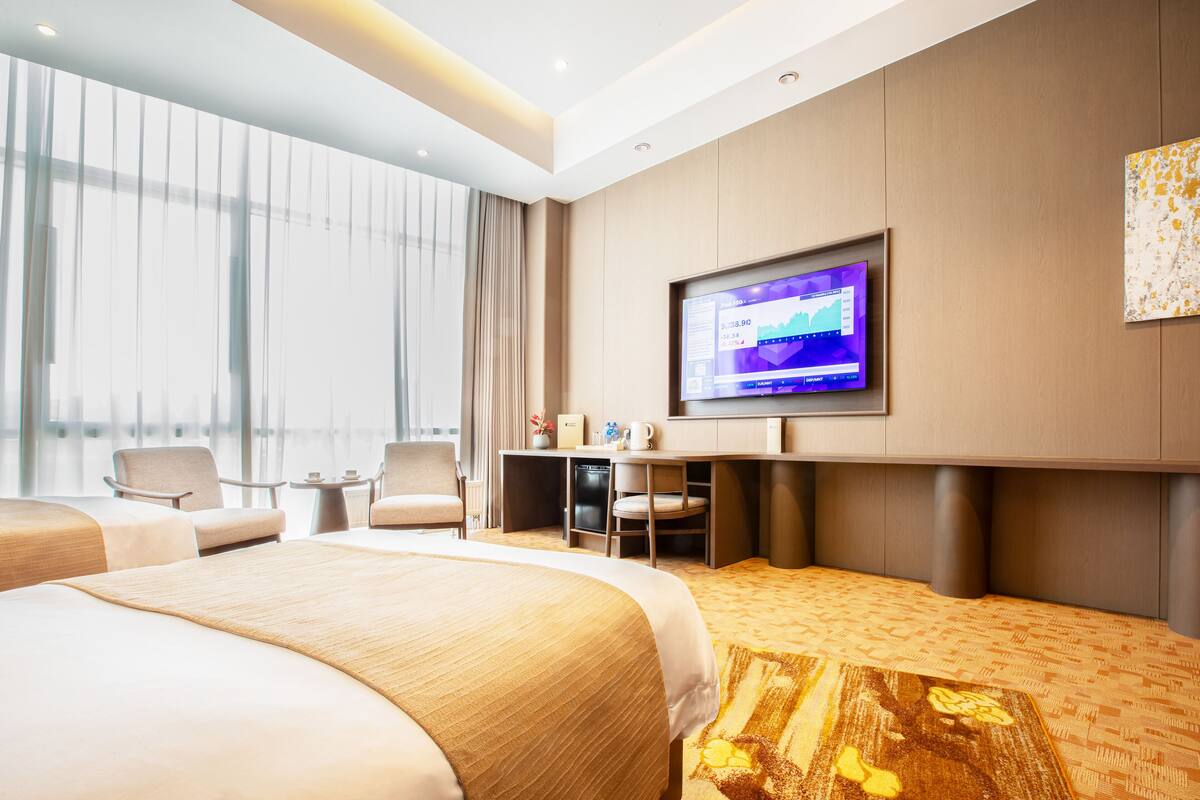 A hotel room with a large bed, a desk with a computer, a television, and a view of the city through large windows.