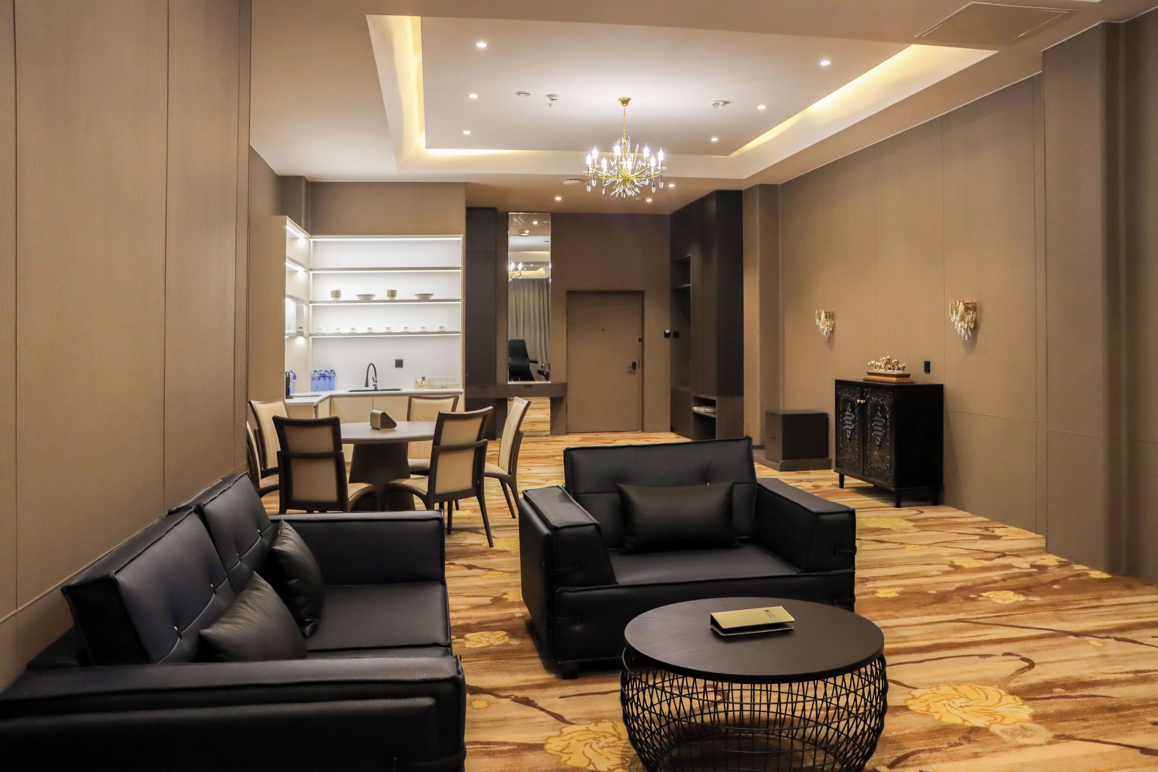 A modern hotel lobby with leather sofas, a round coffee table, a chandelier, and a dining area in the background.