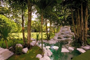 A serene garden with a small waterfall, lush greenery, and a clear pond. - Sheva Om (Pak Kret)