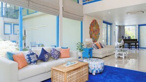 A modern living room with a white sofa, blue and orange cushions, a wooden coffee table, and a colorful wall art piece. - Sheva Om (Pak Kret)