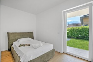 Une chambre à coucher avec un lit, du linge de lit blanc et une vue sur un jardin verdoyant, visible à travers une porte ouverte.