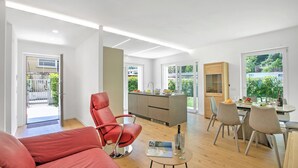 A modern living room with a red chair, a dining area with a table and chairs, a kitchen with a central island, and large windows offering a view of greenery. - Casa Rudolfo Apartment With Private Garden (Riva del Garda)