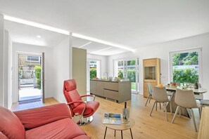 A modern living room with a red chair, a dining area with a table and chairs, a kitchen with a central island, and large windows offering a view of greenery. - Casa Rudolfo Apartment With Private Garden (Riva del Garda)