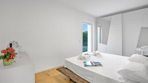 A modern hotel room with a large bed, a bedside table with a vase of flowers, a window with a view of greenery, and a mirror on the wall. - Casa Rudolfo Apartment With Private Garden (Riva del Garda)