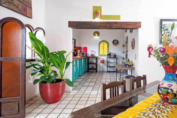 Un comedor con una mesa de madera, sillas y un mantel amarillo brillante. Hay una planta grande en maceta y un florero lleno de flores sobre la mesa.