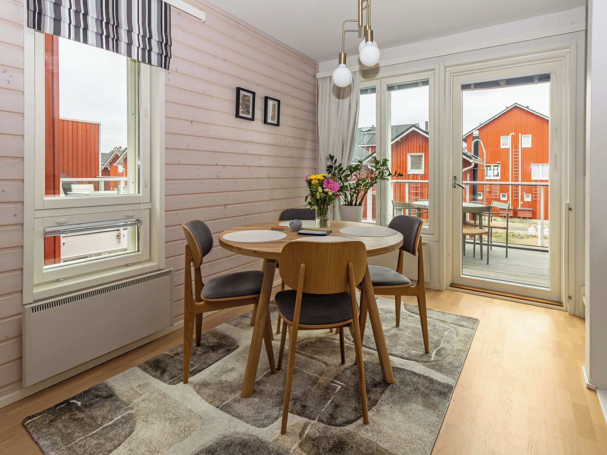 A dining area with a round table, four chairs, a vase with flowers, and a view of red buildings through large windows.