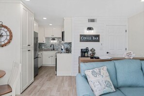 Une cuisine moderne avec des meubles blancs, un panneau indiquant un coin café, et un canapé bleu dans l’espace salon.