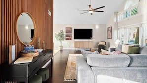 Una sala de estar moderna con una pared de acento de madera, un espejo redondo grande, un ventilador de techo, un sofá gris y un televisor de pantalla plana.