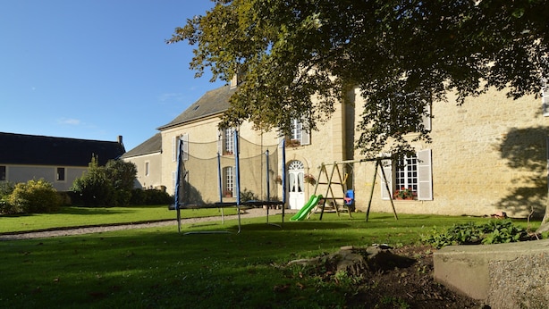 Une zone herbeuse avec une balançoire et un toboggan, un bâtiment en pierre doté de fenêtres, et un arbre qui offre de l’ombre.
