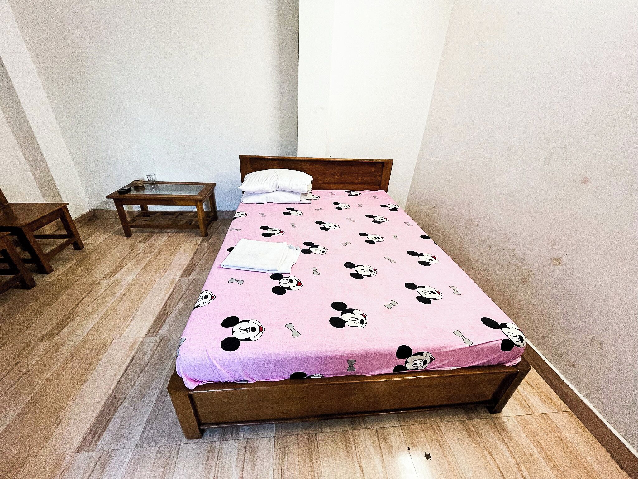 Una cama pequeña y ordenada con una colcha temática rosa de Mickey Mouse, una mesita de noche de madera con una lámpara y una silla de madera.