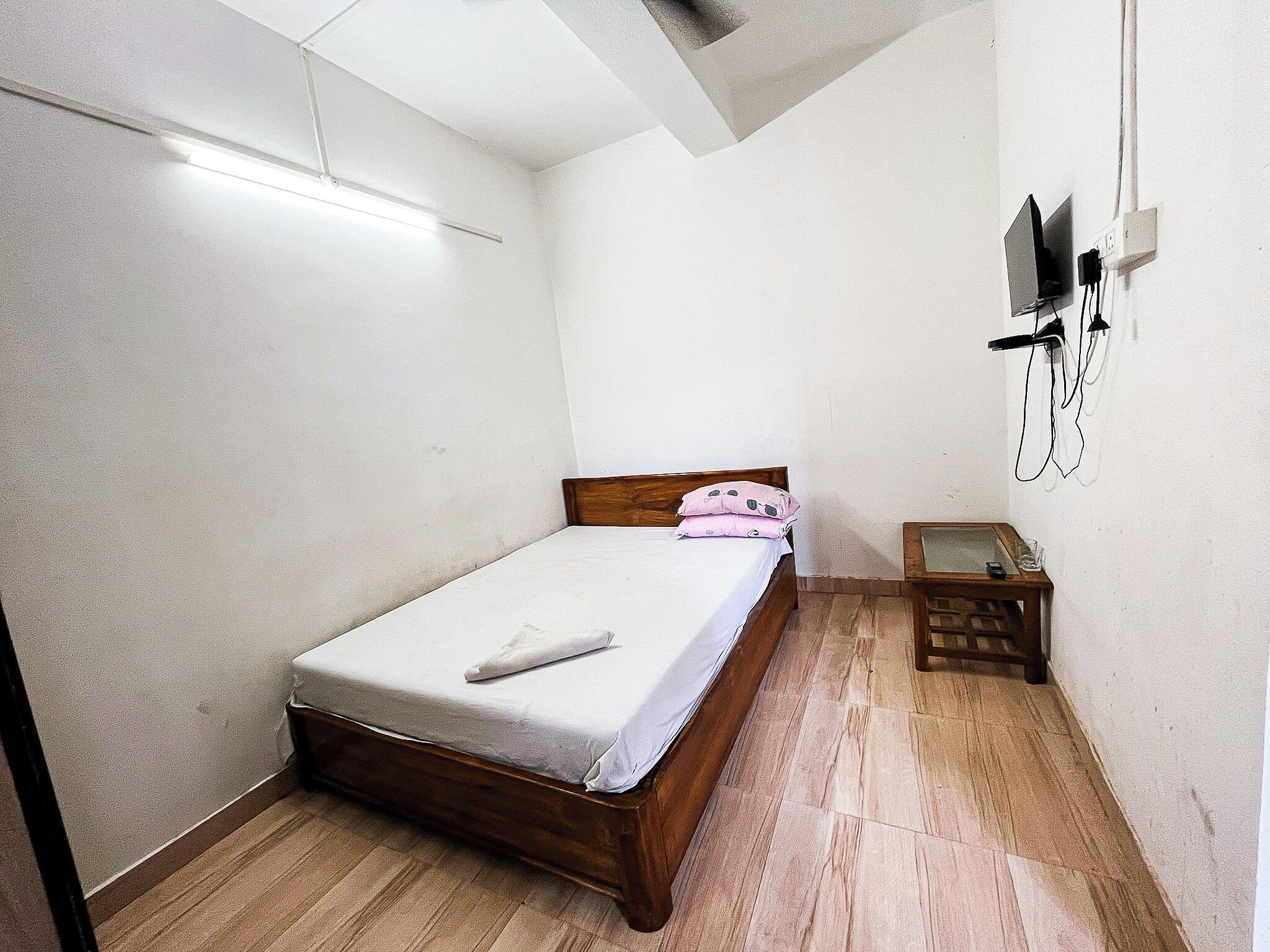 A bedroom with a wooden bed, white walls, a small bedside table, and a wall-mounted TV.
