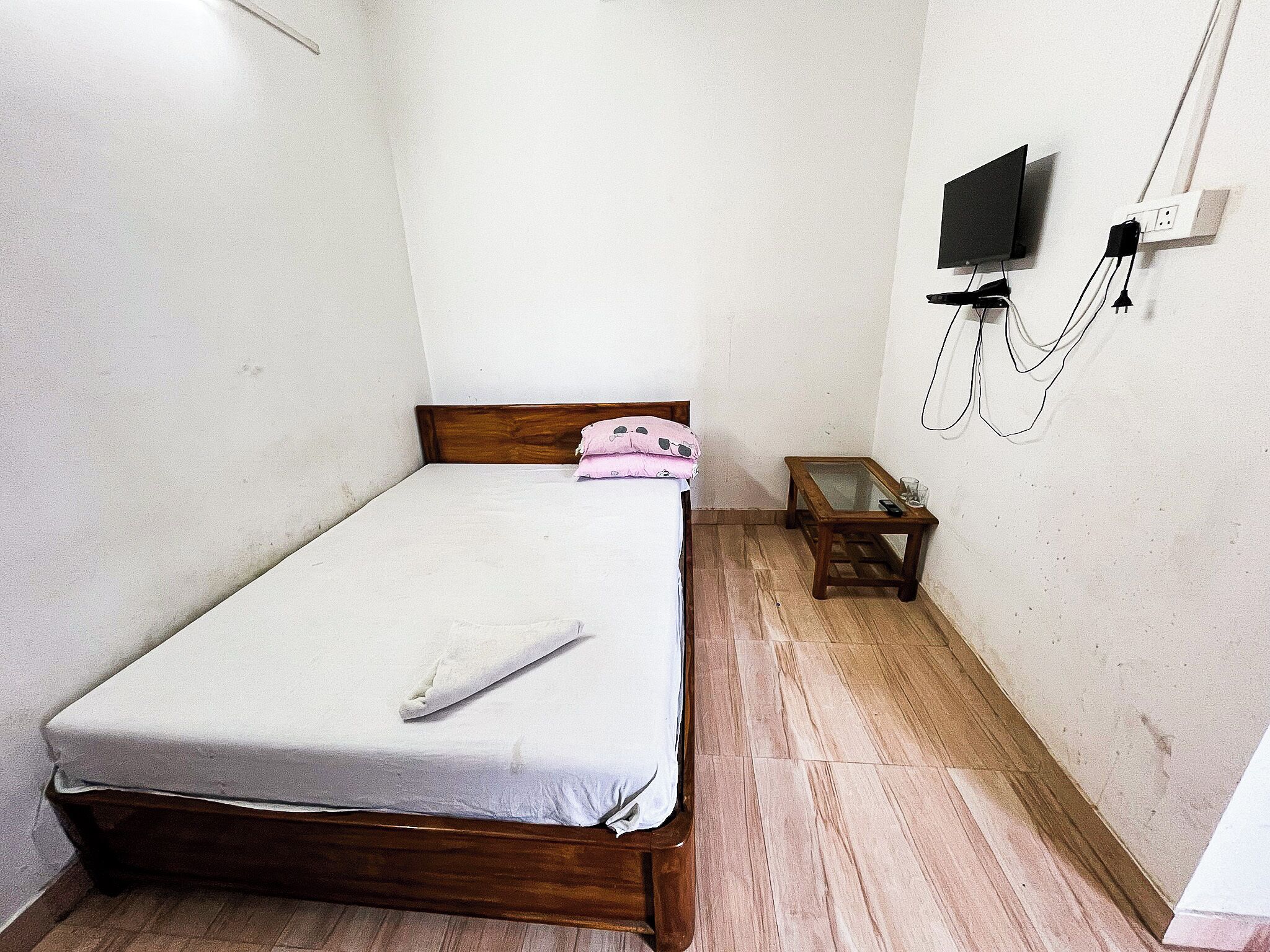 A small, simple room with a single bed, a small table, and a wall-mounted TV.