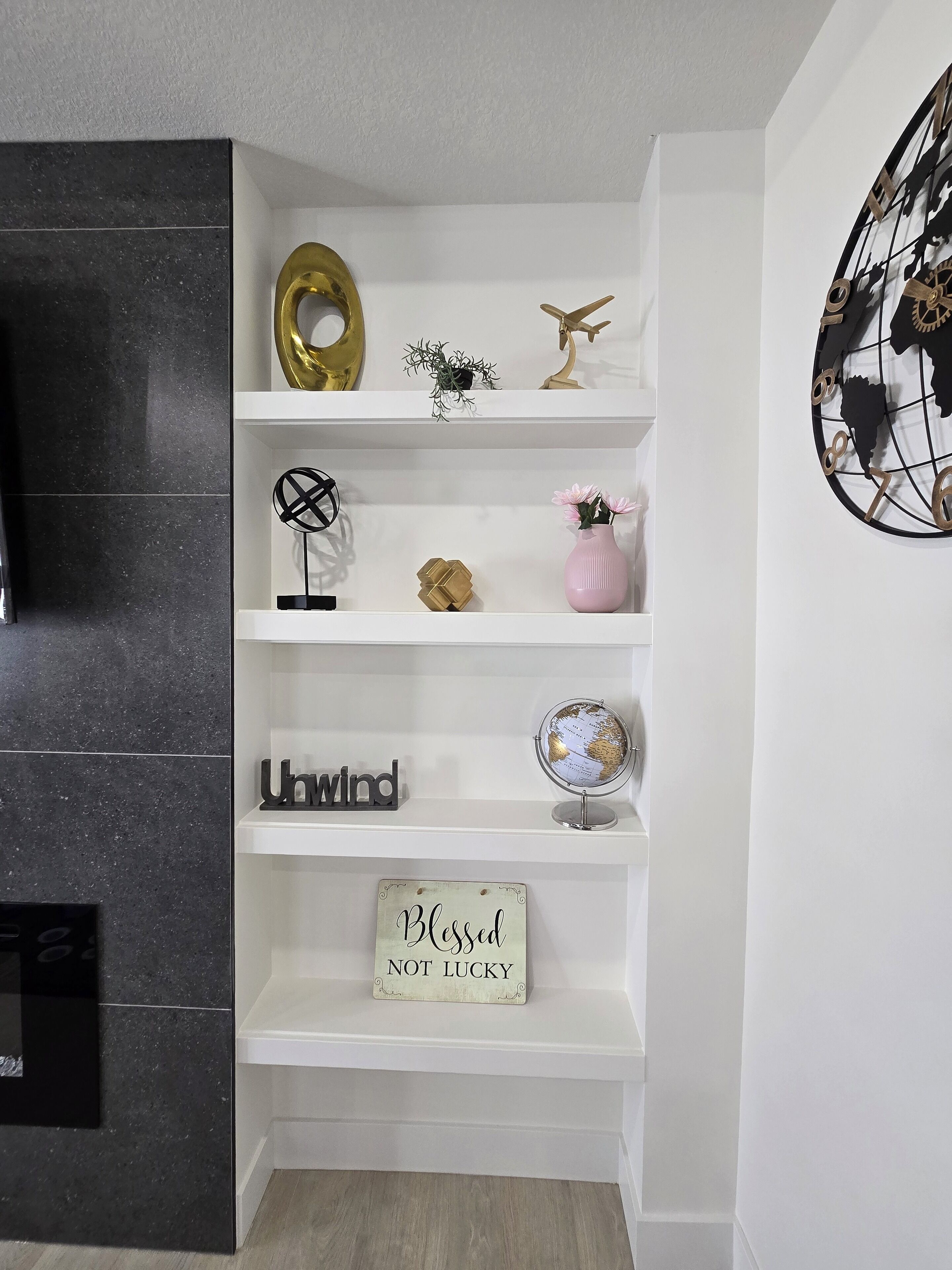 Een witte plank met decoratieve objecten, waaronder een wereldbol, een gouden letter 'H', een klein vliegtuigje, een zwart abstracte sculptuur, een roze vaas met bloemen, en een bordje met de tekst 'Blessed Not Lucky'.