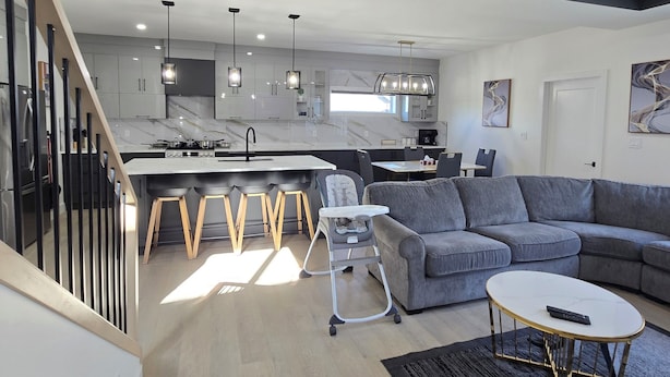 Una sala de estar moderna con un sofá gris, una mesa de centro y una trona para bebé. Se ve una escalera que sube a un nivel superior.